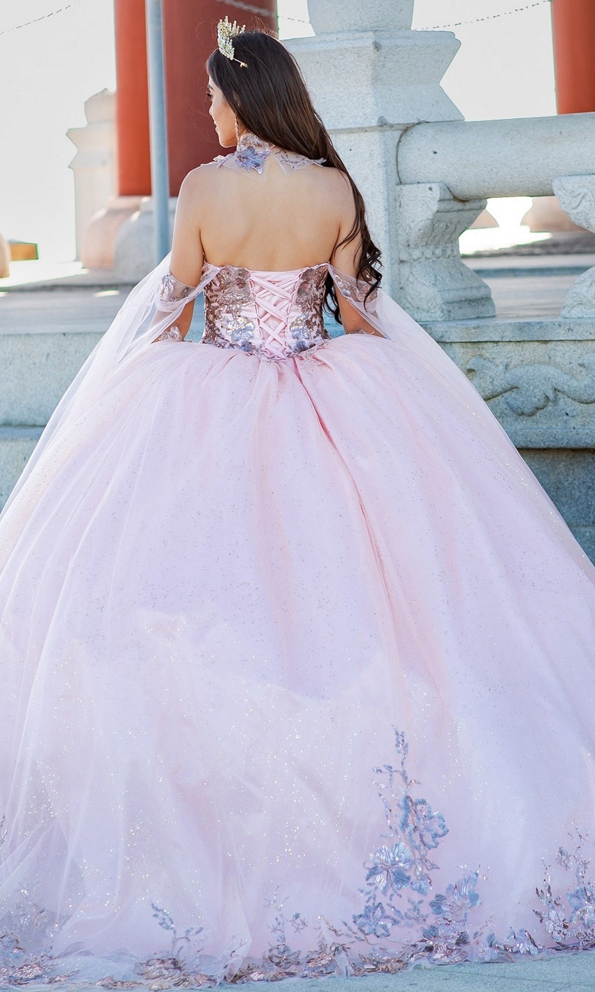 Wearing the Quinceanera Ball Gown 8064J with pink lace embroidery and silver details, a woman with a tiara stands outdoors by stone railings and pillars, facing away from the camera.